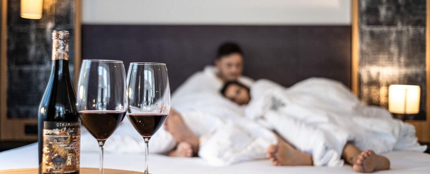 Zwei Gläser Rotwein vor einem kuschelnden Paar im Hotelbett