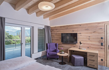 Helles Hotelzimmer mit lila Sessel, Schreibtisch und Balkonblick auf Berge und grüne Landschaft